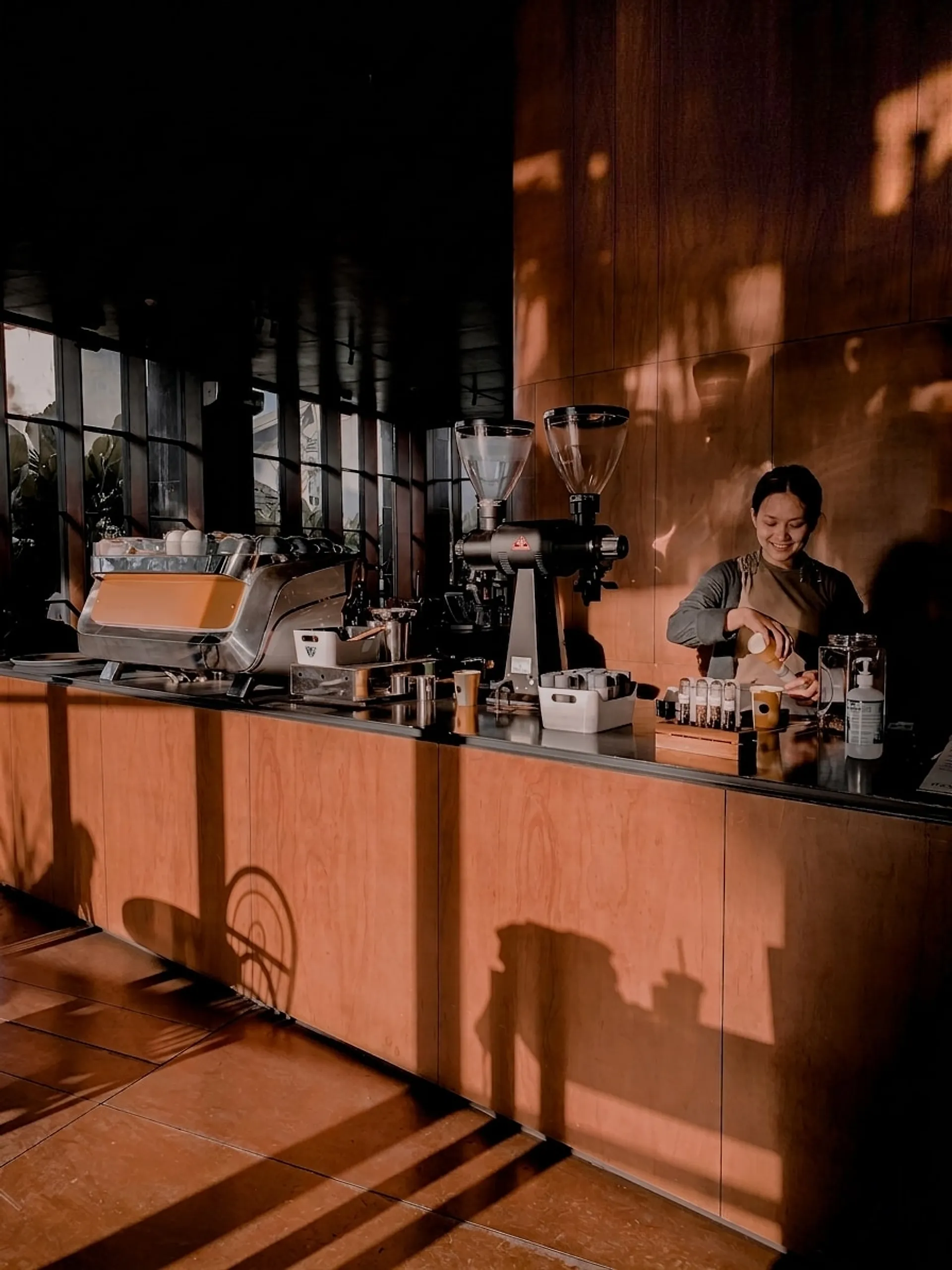 Barista working behind a plywood counter, espresso machine and grinders in low afternoon light